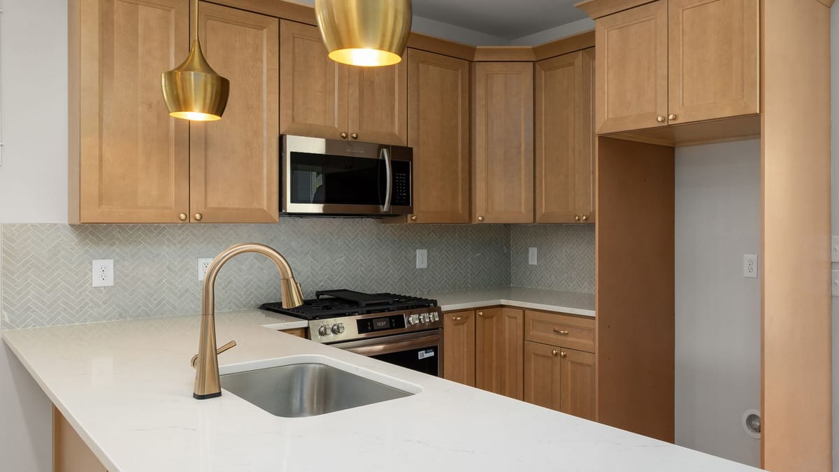 Medium-stained Shaker kitchen cabinets with white counters, brass pendant lights, and a gold faucet